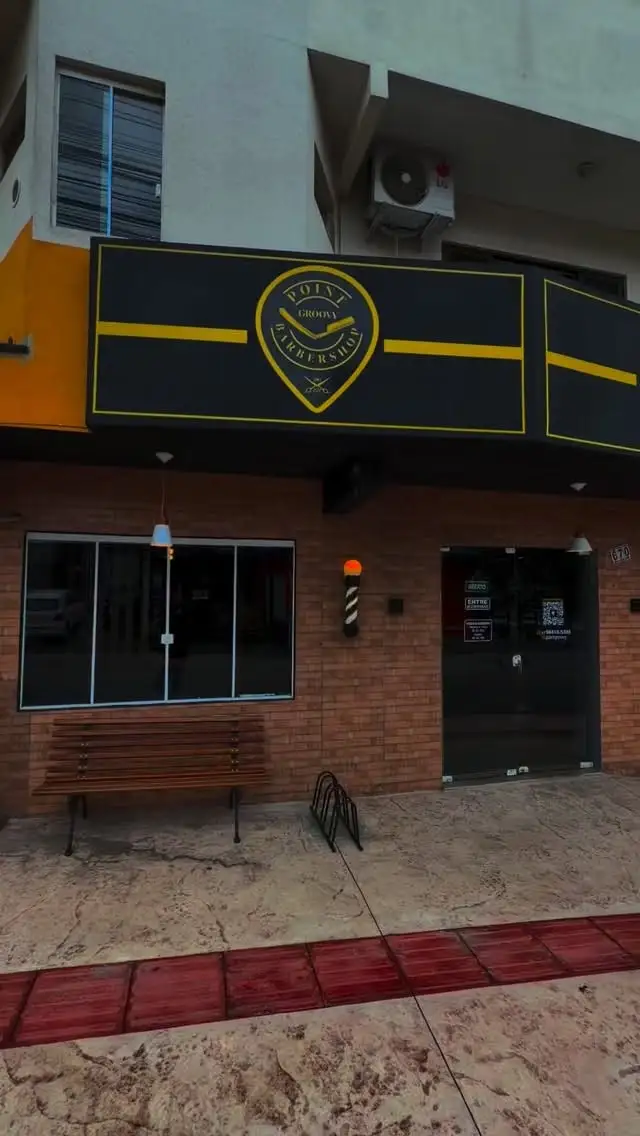 Fachada da Point Groovy em cor: tijolo aparente, letreiro preto com faixa amarela, banco de madeira e poste de barbeiro